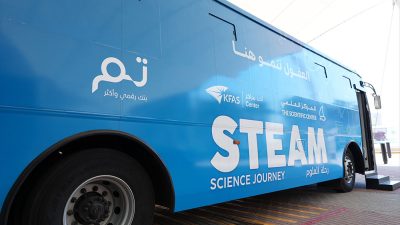 A blue coach parked on paving with its door open, lettered STEAM Science Journey in English and Arabic alongside the logos of KFAS, The Scientific Center and Tam.
