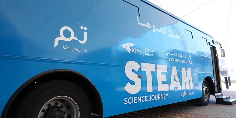 A blue coach parked on paving with its door open, lettered STEAM Science Journey in English and Arabic alongside the logos of KFAS, The Scientific Center and Tam.