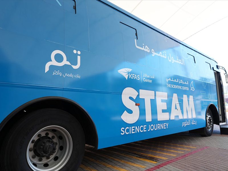 A blue coach parked on paving with its door open, lettered STEAM Science Journey in English and Arabic alongside the logos of KFAS, The Scientific Center and Tam.