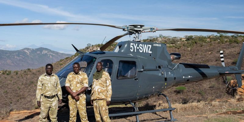 Three rangers in fatigues stand in front of a grey Airbus helicopter registered 5Y-SWZ on a hilltop, with dry hills behind them.