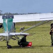 A light aircraft of the Sheldrick Wildlife Trust Aerial Unit stands on a grass airstrip in Kenya with rangers and a vehicle beside it, and a zebra grazing behind the wing.