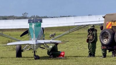 A light aircraft of the Sheldrick Wildlife Trust Aerial Unit stands on a grass airstrip in Kenya with rangers and a vehicle beside it, and a zebra grazing behind the wing.