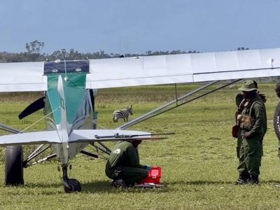 A light aircraft of the Sheldrick Wildlife Trust Aerial Unit stands on a grass airstrip in Kenya with rangers and a vehicle beside it, and a zebra grazing behind the wing.