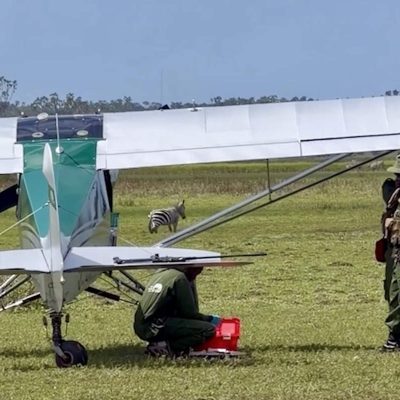 A light aircraft of the Sheldrick Wildlife Trust Aerial Unit stands on a grass airstrip in Kenya with rangers and a vehicle beside it, and a zebra grazing behind the wing.