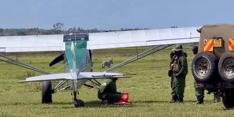 A light aircraft of the Sheldrick Wildlife Trust Aerial Unit stands on a grass airstrip in Kenya with rangers and a vehicle beside it, and a zebra grazing behind the wing.