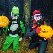 Two SCUBA divers in costume kneel on the gravel bottom of an aquarium exhibit, each holding up a carved jack-o'-lantern, one in a green dinosaur suit and hood and the other in a red and blue suit with a red hood, bubbles rising around them.