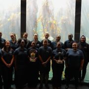 Aquarium staff in dark uniforms stand in front of a tall kelp forest exhibit, two of them holding trophies.