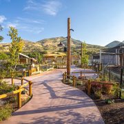 A curving paved path runs through the Wild Utah habitat between timber fencing and young trees, with a tall mesh aviary on the right and dry foothills rising behind.