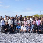 A large group of teenage students and aquarium staff stand and crouch together on a shell-strewn beach on South Africa’s West Coast.