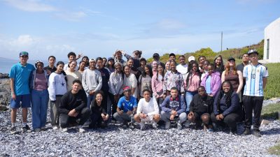 A large group of teenage students and aquarium staff stand and crouch together on a shell-strewn beach on South Africa’s West Coast.