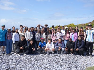 A large group of teenage students and aquarium staff stand and crouch together on a shell-strewn beach on South Africa’s West Coast.