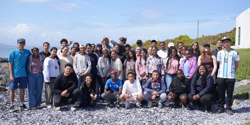 A large group of teenage students and aquarium staff stand and crouch together on a shell-strewn beach on South Africa’s West Coast.