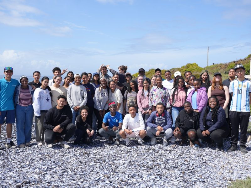A large group of teenage students and aquarium staff stand and crouch together on a shell-strewn beach on South Africa’s West Coast.